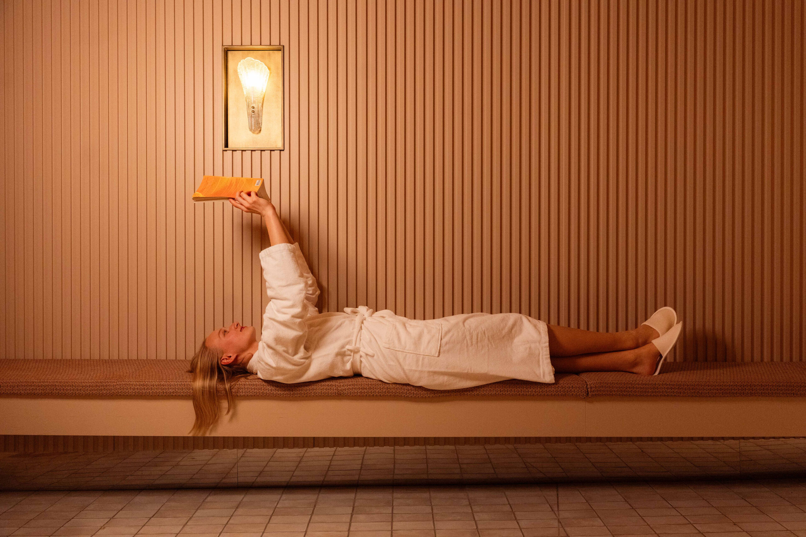 Person lying on a bench in a warm room, reading an orange book under a wall light in a sauna-like space.